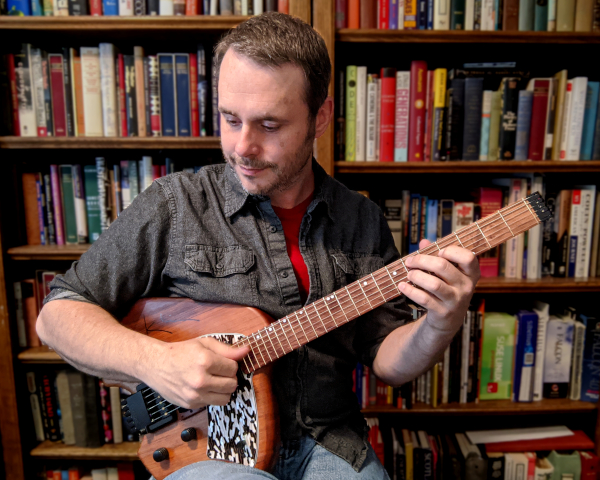 Vic Dillahay playing his guitar at home, in front of bookshelves
